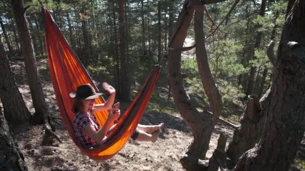 快乐快乐的女人坐在阳光明媚的松树林中的游客吊床上 戴着毛毡帽 在电视里打了个电话 女孩单人游人使用智能手机在网上呆在大自然里 — 图库视频影像