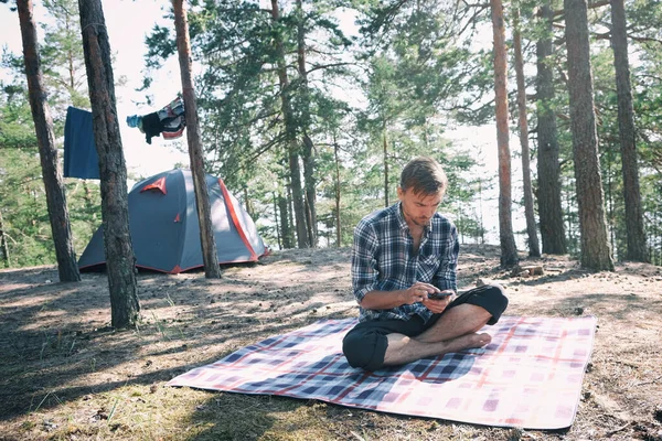 キャンプテントの近くの森の中で毛布の上に座っている男性観光客やスマートフォンを使ってインターネットを接続しようとします モバイルデバイスでのハイキングやアウトドア活動 ロイヤリティフリーのストック画像