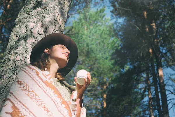 ウールの毛布に包まれた帽子に包まれた穏やかな女性旅行者は 森の中に立って木にもたれかかり マグカップから熱いお茶を飲みます マインドフル リラックスした屋外で あなた自身を扱う ストックフォト