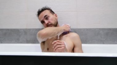 Genç yakışıklı beyaz adam banyoda oturuyor ve vücudu süngerle köpükle yıkıyor. Sportif sakallı adam banyoda duş alıyor. Sabah rutini, vücut bakımı, hijyen konsepti.