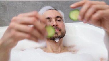 Yakışıklı sakallı adam köpükle banyo yapıyor, salatalık maskesi takıyor. Ev banyosunda spa tedavisi. İnsan sağlığı, vücut bakımı ve kendini sevme kavramı..