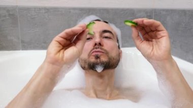 Komik yakışıklı sakallı adam salatalık maskesiyle köpük banyosunda rahatlıyor, salatalık yiyor. Ev banyosunda spa tedavisi. İnsan sağlığı, vücut bakımı ve kendini sevme kavramı..