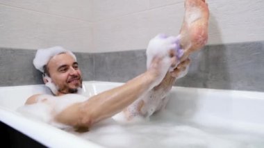 Genç yakışıklı beyaz adam köpükle banyoya uzanıyor ve vücudu sünger pufla yıkıyor. Erkek sağlığı, vücut bakımı, hijyen konsepti..