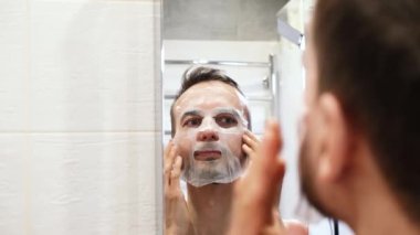 Aynanın yansıması gülümseyen bin yıllık adam banyoda kozmetik maske takıyor. Evde kişisel bakım, sağlık ve kaplıca tedavisi.