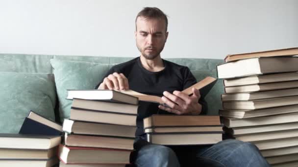 若いひげを生やした男のスピードリーダー自宅でソファに座って すべての側面に本の山に囲まれ 深く読書を夢中にさせた 学習と教育 情報の検索 — ストック動画