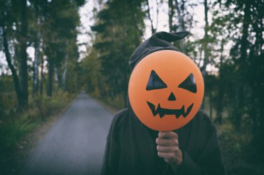 Keşiş ya da büyücü kostümlü gizemli adam sonbahar yolunda tek başına dikilirken yüzünde korkunç balkabağı resmi olan turuncu hava balonu tutuyor. Cadılar Bayramı konsepti