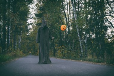 Keşiş ya da büyücü kostümlü gizemli bir adam sonbahar yolunda tek başına duran balonlu turuncu balon ve balkabağı kafalıyı tutuyor. Cadılar Bayramı konsepti.