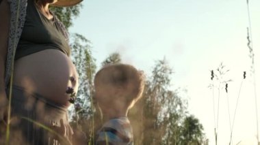 Bebek bekleyen otantik aile anları. Küçük çocuk hamile annesine sarılıyor ve doğada karnını öpüyor. Hamile kadın yaz sahasında gün batımında dışarıda.