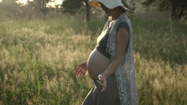 Gün batımında yeşil yaz sahasında yürüyen boho tarzı elbiseli hamile bir kadın. Bebek beklemenin otantik anları.