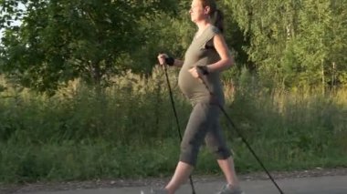 Hamilelikte boş zaman. Hamile kadın yaz parkında yürüyüş direğiyle İskandinav yürüyüşü yapıyor..