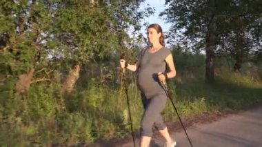 Kamera, günbatımında kuzey parkında yürüyen ve gülümseyen hamile bir kadını takip ediyor. Bekleyen sağlıklı anne yürüyüş direğiyle dışarıda antrenman yapıyor..