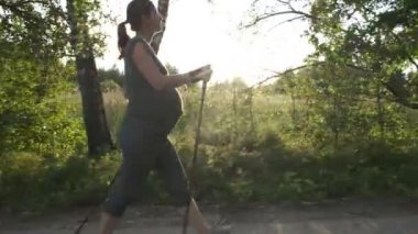 Tam boy yan görüş aktif hamile kadın dışarıda yürüyüş direkleri ile kuzeye yürüme pratiği yapıyor. Bekleyen sağlıklı anne gün batımında yaz parkında antrenman yapıyor..