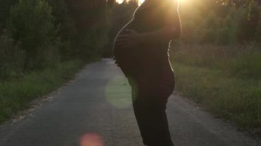 Gün batımında kırsal yolda karnını okşayan hamile bir kadın. Annelik, bebek beklentisi, mutlu hamilelik konsepti..