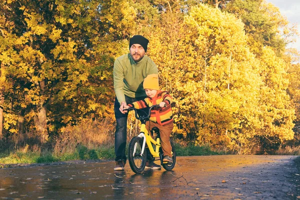 父は子供に秋の公園で自転車に乗るように教えています 子供はサイクリングを練習する 幸せな家族の瞬間 父と息子との時間 候補者のライフスタイル写真 — ストック写真