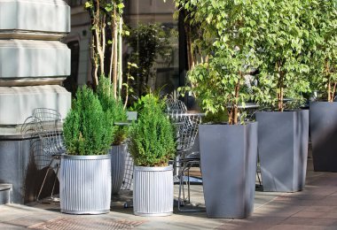 Yapay selvi ağaçlarının gölgesinde bir kafenin masa ve sandalyeleri.