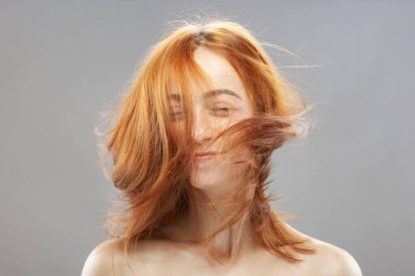 Güzel kızıl saçlı kız, el sallayan saçlarıyla poz veriyor. Gri arkaplanda stüdyo resmi.