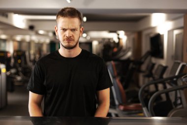 Kızgın adam dikilip kameraya bakıyor. Odaklanamayan bir spor salonunda.