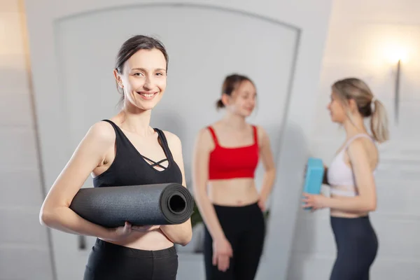 Yoga instructor posing with yoga mat. Two girls chatting in the background.