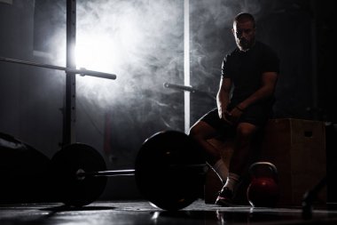 Ağırlık kaldırma ekipmanıyla içeride poz veren kaslı bir sporcunun silüeti. Duman arkaplanı.