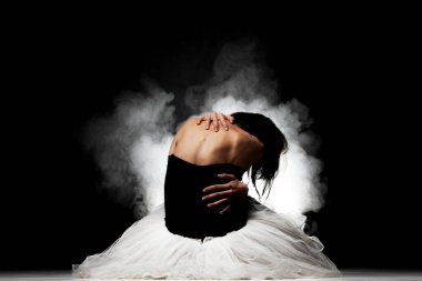 Balerin elinde etekle yerde oturuyor, arkasında dumanla poz veriyor.