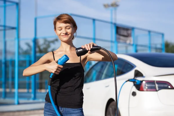 Piękna Młoda Dziewczyna Obok Elektrycznego Samochodu Pomieszany Dwoma Kablami Ładującymi — Zdjęcie stockowe