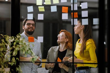 Three people brainstorm ideas for a project using sticky notes on a glass wall in an office. Natural light fills the space, and they share thoughts enthusiastically.