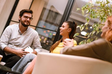 Three friends enjoy a warm conversation in a stylish indoor space filled with plants and soft lighting, sharing laughter and friendly vibes in the evening.