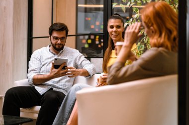 Three friends are seated in a stylish indoor area. One is using a tablet, while the others chat and sip drinks, creating a relaxed and friendly atmosphere together.