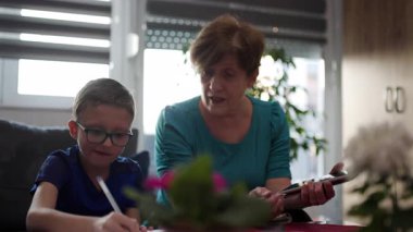 Grandmother helps grandson with homework. They are sitting at a table, talking and smiling.