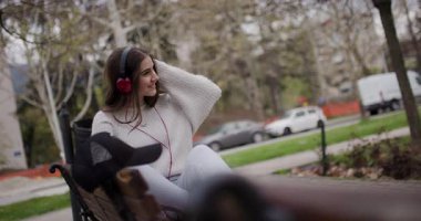 Teenage Girl Listens to Music on Red Headphones While Sitting on Park Bench
