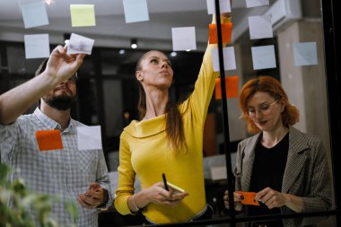 In a vibrant office, three colleagues engage in a brainstorming session, using colorful sticky notes on a glass wall to share ideas and enhance collaboration.