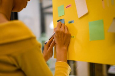 A woman is writing on a large yellow poster covered with colorful sticky notes in a bright, open office. The atmosphere is energetic, capturing the spirit of teamwork and idea generation.