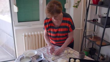 An elderly woman is making bread in her kitchen. She rolls dough balls and dips them into oil before placing them on a baking tray.