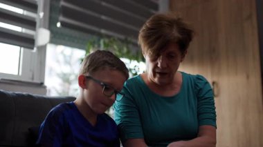 Grandmother and grandson spend quality time together, reading and talking. The boy wears glasses and sits next to his grandmother on a sofa.
