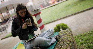 Teenage Girl Using Her Smartphone and Taking Notes in a Park While Sitting on a Blanket