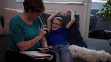 Grandmother and grandson using a phone and notepad, talking. A small white dog sits on the couch between them.