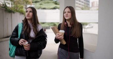 Two Teenage Girls Walk and Talk Between Classes at School, Drinking Coffee and Discussing Homework