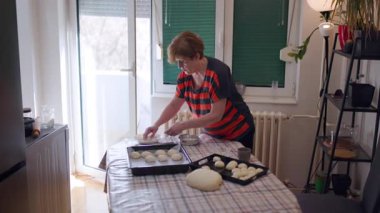 An elderly woman skillfully prepares dough and arranges it on baking trays, showcasing the art of homemade baking in her cozy kitchen.
