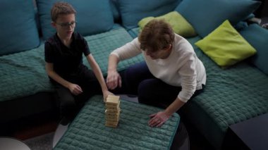 Grandmother and grandson are playing a game of blocks on the living room sofa. The child is carefully extracting a wooden block from the structure.