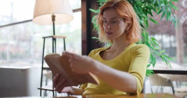 Kahve dükkanındaki bir masada, kızıl saçlı bir kadın kitap okuyor. O, büyük bir pencerenin yanında oturur ve dışarıya bakar. Yağmur yağıyor gibi görünüyor..