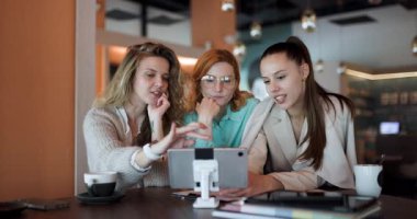 Üç kadın modaya uygun bir kafede toplanmış, stratejileri tartışıyor ve dijital bir ekrana odaklanırken içgörülerini paylaşıyorlar..