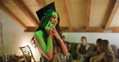 Heyecanlı mezun kutlama sırasında telefonda harika haberleri paylaşıyor. Arkadaşlar mezuniyet şapkasıyla ev partisinin arka planında toplandılar..