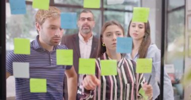 Kafkas iş adamlarından oluşan bir ekip cam pencerenin yanında yapışkan notlarla bekliyor. Renkli çıkartmalı Scrum metodolojisi. İş arkadaşları proje fikirlerini tartışıyor. 50 fps 'de kaydedildi.