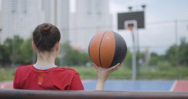 Kırmızı formalı bir bayan basketbolcu, şehir dışındaki bir sahada elinde bir topla bankta oturur, yansıtır ve rahatlatır..