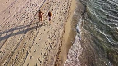 Güneşli bir günde, okyanus dalgaları yakınında kumlu bir plaj boyunca yürüyen iki genç kadın. Şnorkel malzemeleri taşıyorlar, kaygısız yaz tatillerinin tadını çıkarıyorlar..