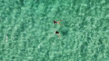 İki kadın güzel, berrak turkuaz suda yüzüyor ve dinleniyorlar. Arkadaşların kumsaldaki yaz tatilinin tadını çıkardıkları hava aracı görüntüsü..