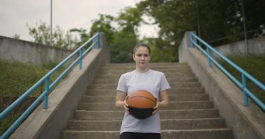 Genç bir bayan basketbolcu, dinamik ve kentsel bir ortamda becerilerini ve atletizmini sergileyerek topunu açık havada sallıyor..