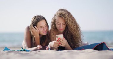 Akıllı telefonu inceleyen ve kumsalda dinlenirken içeriğini paylaşan iki genç kızın gündelik ve neşeli etkileşimi..