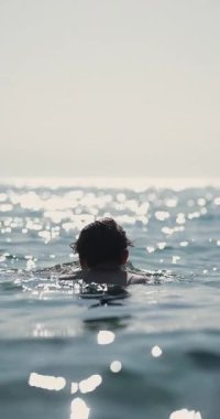 Denizde yüzen bir adamı gösteren sakin bir görüntü, arkadan bakıldığında, suda parlayan bir güneş. Sinema kamerasında 120 fps 'de kaydedildi..