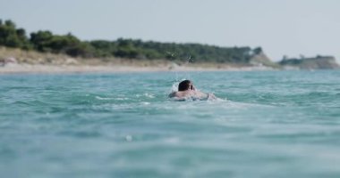 Denizde yüzen bir adamın canlı mavi suları ve arka planda manzaralı bir kıyı şeridini gösteren yakın plan fotoğrafı. Sinema kamerasında 120 fps 'de kaydedildi..
